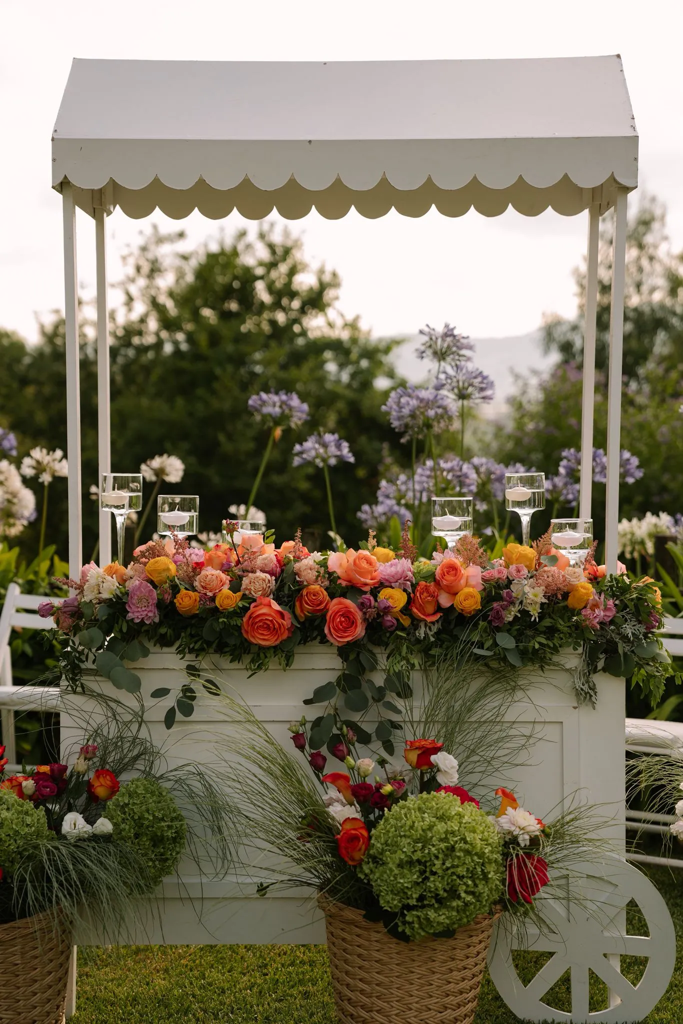 A white scalloped drinks cart overflowing with seasonal flowers