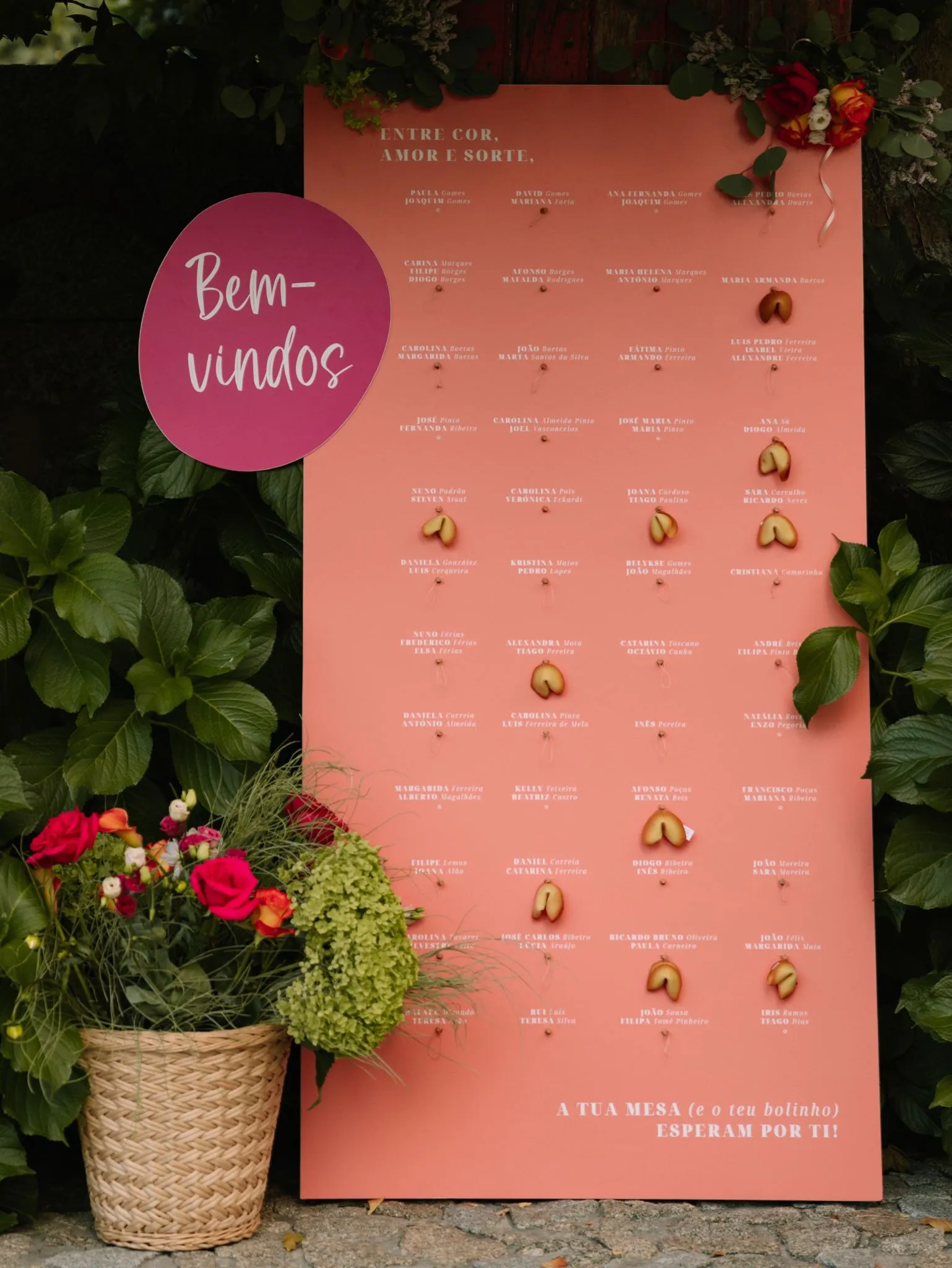 A coral 'Bem-vindos' seating chart framed by greenery